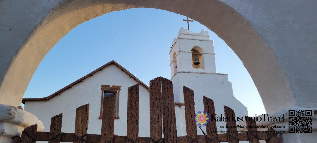 La fachada de una iglesia de adobe blanca en San Pedro de Atacama, enmarcada por un arco de adobe en primer plano.